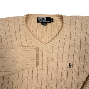 May include: A beige V-neck cable knit sweater featuring a dark brown embroidered polo player logo on the chest. The interior neck label reads Polo by Ralph Lauren with a size L tag. The garment is constructed with a textured vertical cable pattern.