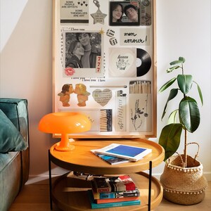 Op de afbeelding: Een houten collage met foto's, een vinylplaat en decoratieve items. Een feloranje paddenstoellamp staat op een houten bijzettafel met twee niveaus. Een potplant in een geweven mand staat naast de tafel.