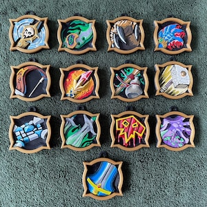 May include: Thirteen circular, tan-framed pendants featuring colorful, raised relief illustrations of fantasy-themed items, including a skull, a glowing crystal, a hammer, swords, and various magical symbols, arranged in a grid on a textured green surface.