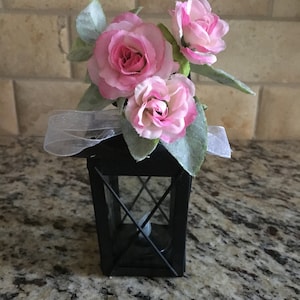 May include: A small black lantern with a white ribbon and pink silk flowers on top. The lantern has a clear glass window and a candle inside.