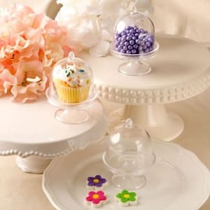 Può includere: Tre supporti per torte in plastica trasparente con coperchi a cupola. I supporti sono di dimensioni diverse e sono esposti su una superficie bianca. Il supporto più piccolo contiene codette viola all'interno, il supporto intermedio contiene un cupcake giallo con glassa bianca e codette, e il supporto più grande è vuoto.