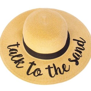 May include: A straw sun hat with a black ribbon and the text "talk to the sand" embroidered in black.