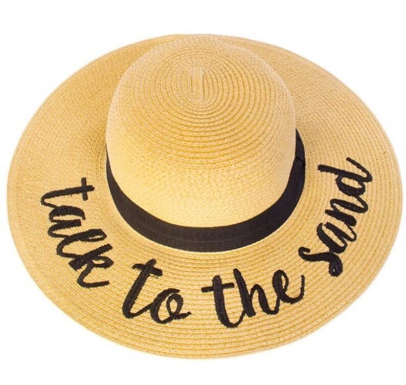 May include: A straw sun hat with a black ribbon and the text "talk to the sand" embroidered in black.