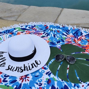 May include: White sun hat with black trim and the text "hello sunshine" embroidered on the brim. The hat is lying on a colorful round beach towel with a green leaf and sunglasses.