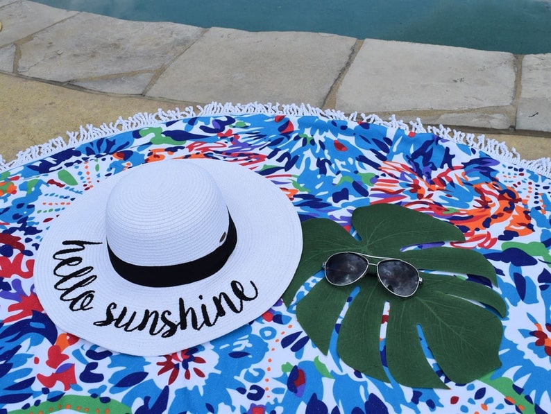 May include: White sun hat with black trim and the text "hello sunshine" embroidered on the brim. The hat is lying on a colorful round beach towel with a green leaf and sunglasses.