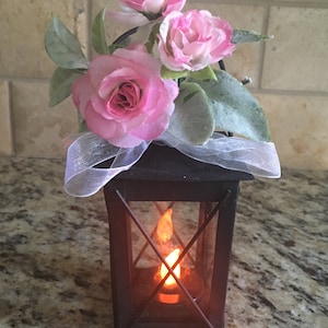 May include: A black metal lantern with a glass window and a lit candle inside. The lantern is decorated with pink silk flowers and a white ribbon.