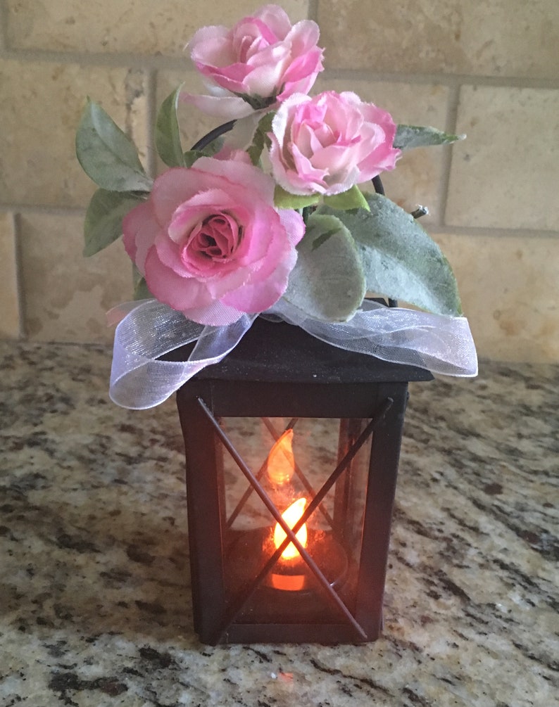 May include: A black metal lantern with a glass window and a lit candle inside. The lantern is decorated with pink silk flowers and a white ribbon.