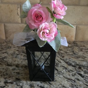 May include: A small black lantern with a white ribbon and pink silk flowers on top. The lantern has a clear glass window and a candle inside.