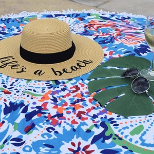 May include: A straw hat with a black band and the text "life's a beach" on a colorful patterned beach towel. A glass of white wine and sunglasses are on a green leaf.