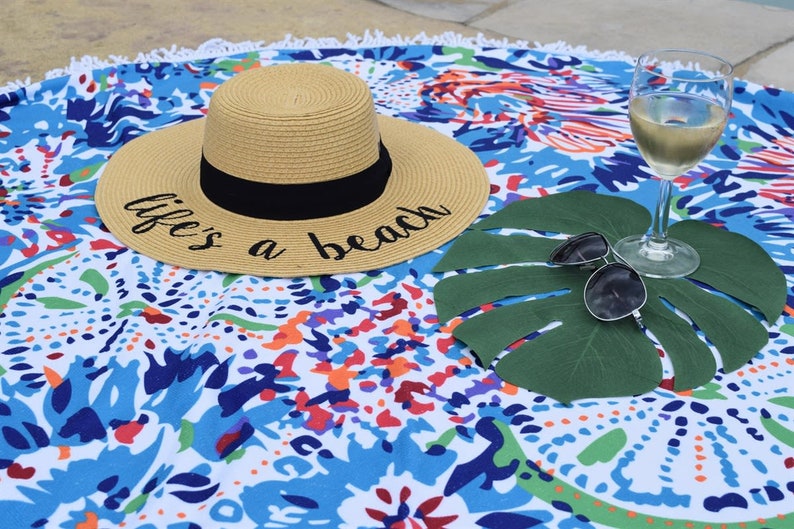 May include: A straw hat with a black band and the text "life's a beach" on a colorful patterned beach towel. A glass of white wine and sunglasses are on a green leaf.