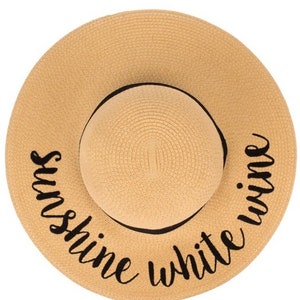 May include: A tan straw sun hat with a black ribbon and the text "Sunshine White Wine" embroidered on the brim.