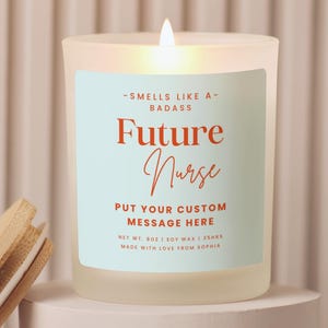 May include: A lit, frosted glass candle with a light blue label that reads "Future Nurse." The candle sits on a white pedestal, with a wooden lid beside it. The candle is made with soy wax and has a burn time of 35 hours.