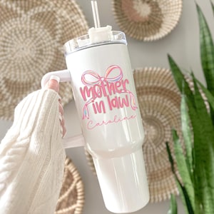 May include: White tumbler with a handle, a clear lid, and a straw. The tumbler features the text "Mother in Law" in pink and a bow design. The name "Caroline" is written below the text in pink.