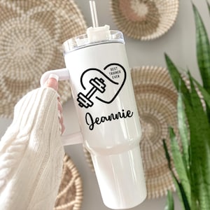 May include: White tumbler with a handle, straw, and lid. The tumbler features a black heart design with a dumbbell and the text "BEST TRAINER EVER" and the name "Jeannie". The tumbler is designed for cold beverages.