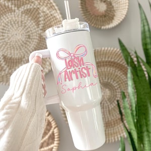 May include: A white tumbler with a handle and a clear lid. The tumbler features the text "Lash Artist Sophia" in pink and a colorful bow design. A clear straw is inserted into the lid.