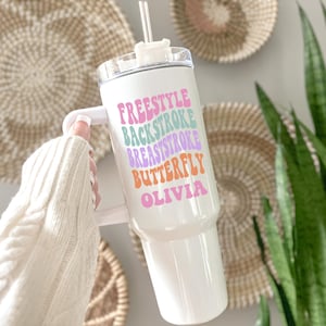 May include: White tumbler with a handle and a clear lid, featuring a straw. The tumbler has the words "FREESTYLE BACKSTROKE BREASTSTROKE BUTTERFLY OLIVIA" in colorful, wavy text. The background includes woven wall decor and greenery.