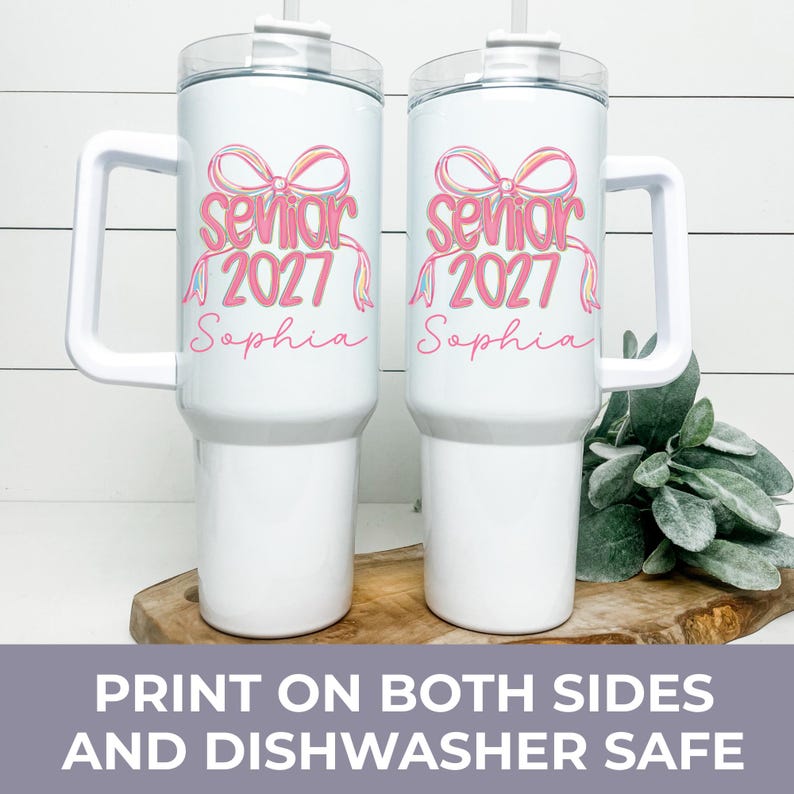 May include: Two white stainless steel tumblers with handles and clear lids. Each features a pink graphic of a bow above the text "senior 2027" and the name "Sophia" in pink script. The design is printed on the front of both insulated cups.