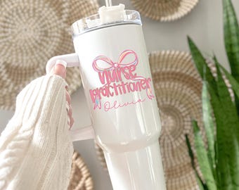May include: White tumbler with a handle, a clear lid, and a straw. The tumbler features the text "Nurse Practitioner Olivia" in pink and blue script with a bow graphic. The tumbler is held by a person wearing a white sweater.