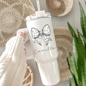 May include: White tumbler with a handle, a clear lid, and a straw. The tumbler features a black and white volleyball bow design and the name "Camilla" in script. The background includes woven wall decor and greenery.