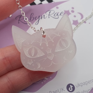 Può includere: Una collana con un ciondolo a forma di gatto bianco traslucido, decorato con un design inciso dettagliato di un viso di gatto, inclusi occhi, naso e baffi. Il ciondolo è su una catena d'argento.