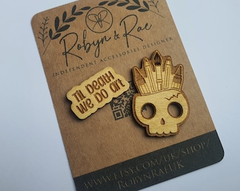 Può includere: Due spille di legno su una carta marrone. Una spilla è un teschio con matite e pennelli, l'altra dice "Til death we do art". La carta ha il testo "Robyn & Rae Independent Accessories Designer".