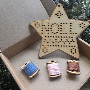 May include: A wooden star-shaped Christmas ornament with the word "NOEL" spelled out in holes, alongside three spools of thread in blue, pink, and brown. The items are in a cardboard box, suggesting a craft or festive theme.