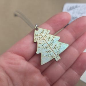May include: A small, glittery Christmas tree ornament. The tree is light green with a detailed brown branch design. It is held by a clear string and has a silver metal loop at the top. The ornament is a festive holiday decoration.