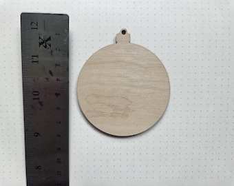 May include: A wooden Christmas ornament in a round shape with a small hole at the top for hanging. The ornament is made of light-colored wood and is approximately 4 inches in diameter. A metal ruler is next to the ornament.