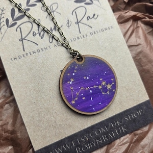 Puede incluir: Un collar con colgante redondo de madera con un diseño celestial púrpura y dorado. El colgante presenta un cielo nocturno pintado con constelaciones y estrellas. El collar tiene una cadena de color bronce y se muestra en una tarjeta con el nombre del diseñador.