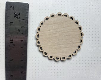 May include: A round, unfinished wooden coaster with a scalloped edge. A metal ruler is positioned next to the coaster, showing measurements in inches. The coaster is made of light-colored wood.