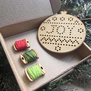 Peut inclure: Un ornement de Noël en bois avec le mot "JOY" découpé, accompagné de trois bobines de fil rouge, vert et vert citron, le tout dans une boîte en carton. L'ornement rond présente un motif décoratif.