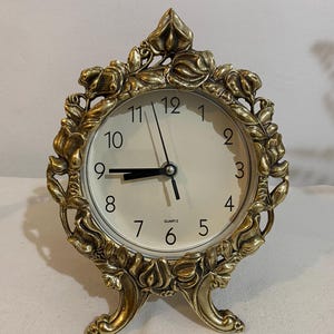 May include: A gold-colored quartz clock with a cream-colored face. The clock has a decorative floral design around the face and on the base. The clock hands point to approximately 8:45. The clock is a decorative home decor item.