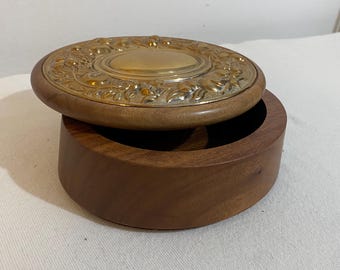 Könnte beinhalten: Eine runde Holzbox mit Deckel. Der Deckel ist mit einer goldfarbenen Metallplatte mit einem verzierten floralen Design verziert. Die Box ist geöffnet und zeigt das Innere. Das Holz hat einen warmen, braunen Farbton.