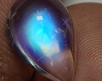 May include: A pear-shaped, polished gemstone with a translucent, iridescent surface. The stone displays a captivating play of color, with a central area of bright blue and surrounding hues of purple and white. The gem is held between fingers.