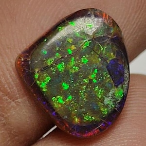 May include: A polished, irregular-shaped dark opal gemstone featuring a vibrant play of color with intense neon green, blue, and red flashes across its surface. The stone displays a distinct dark base tone with a glossy, reflective finish.