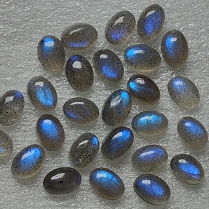 May include: A selection of oval labradorite gemstones. Each stone has a polished surface, displaying a mix of grey and blue tones. The stones exhibit a shimmering, iridescent effect, with flashes of blue light.