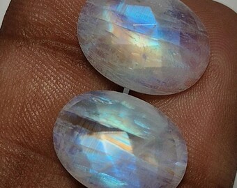 May include: Two oval-shaped moonstone gemstones with iridescent blue, yellow, and orange flashes. The stones are faceted, catching the light and displaying a play of colors. The gemstones are set against a neutral background.