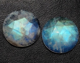 May include: Two round, faceted moonstone gemstones with a blue and white iridescent glow. The gemstones are set against a black, textured background, highlighting their natural beauty and potential for jewelry making.
