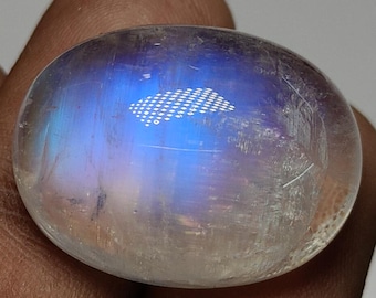 Könnte beinhalten: Ein polierter, oval geformter Mondstein. Der Stein hat ein durchscheinendes Aussehen mit einem blau-weißen schillernden Glanz. Die Oberfläche weist subtile innere Texturen und Lichtreflexionen auf, was auf einen unbehandelten Edelstein hindeutet.