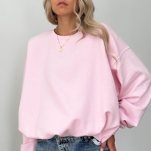 Puede incluir: Una sudadera rosa claro con cuello redondo. La sudadera tiene mangas largas y un ajuste holgado. La modelo lleva vaqueros azules y un collar dorado. La sudadera es de color liso y parece estar hecha de un material suave.
