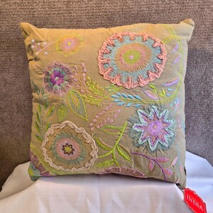 May include: A square, tan decorative pillow with colorful floral embroidery. The embroidered flowers feature shades of pink, blue, green, and purple. The pillow is likely made of a soft fabric and is designed for decorative use.