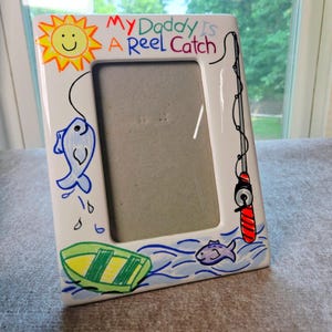 May include: A white ceramic picture frame featuring hand-painted illustrations of a yellow sun, a blue fish, a green boat, and a red and black fishing rod. The text "My Daddy Is A Reel Catch" is written in colorful lettering across the top border.