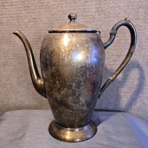 May include: A vintage silver coffee pot with a curved spout and handle. The pot has a lid with a small knob and sits on a round base. The silver surface shows signs of age and use.