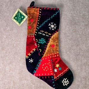 May include: A velvet Christmas stocking featuring a patchwork design of red, black, and brown fabric panels. The stocking is decorated with colorful embroidered stitching, including snowflake and tree motifs. A diamond-shaped tag is attached.