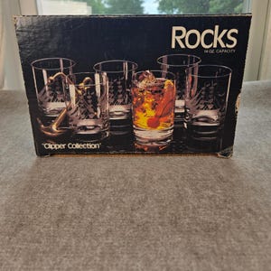 Peut inclure: Une boîte en carton noir pour un ensemble de verres Rocks de la collection Clipper. La boîte présente des images de verres transparents avec des motifs de voiliers gravés et une capacité de 14 oz. Un verre contient un liquide ambré avec une cerise.