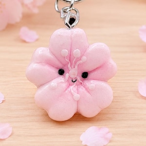 Puede incluir: Un llavero de metal plateado con un cierre en forma de flor de cerezo sakura y un dije colgante de flor esculpida en color rosa claro. El dije tiene un acabado suave y brillante con una pequeña cara pintada a mano con dos ojos negros.
