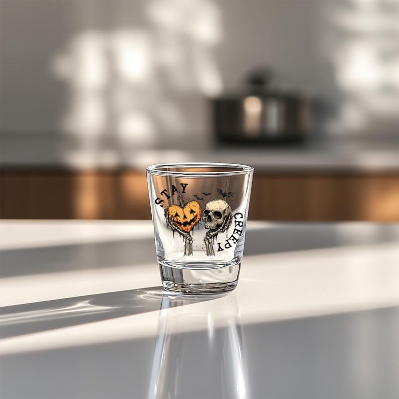 May include: A clear shot glass with a Halloween-themed design. The design features a jack-o'-lantern heart and a skull held by skeletal hands, with the words "STAY CREEPY" printed on the glass. The glass is on a white surface.
