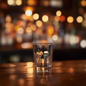 May include: A clear shot glass with a Halloween-themed design. The design features a pumpkin heart and a skull with the words "Stay Creepy" printed on the glass. The shot glass is sitting on a wooden surface.