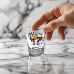 May include: A clear shot glass with a Halloween-themed design. The design features a jack-o'-lantern and a skull with the words "STAY CREEPY" printed on the glass. The shot glass is held by a hand.