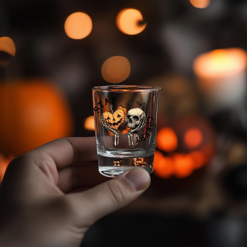 May include: A clear shot glass with a Halloween-themed design. The design features a jack-o'-lantern heart and a skull with the words "Stay Creepy". The glass is held in a hand, with blurred pumpkins and lights in the background.